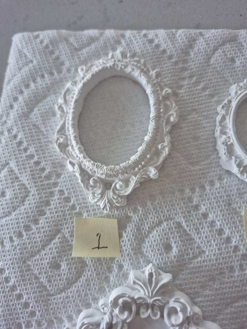 May include: White, ornate oval picture frame with decorative scrollwork. The frame is made of a solid material and is placed on a white, textured surface. A small yellow sticky note with the number "1" is visible.