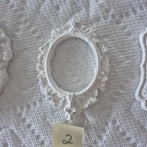 May include: White oval-shaped picture frame with ornate detailing. The frame has a decorative floral design around the edges. A small yellow sticky note with the number "2" is visible below the frame. The frame is on a white textured surface.