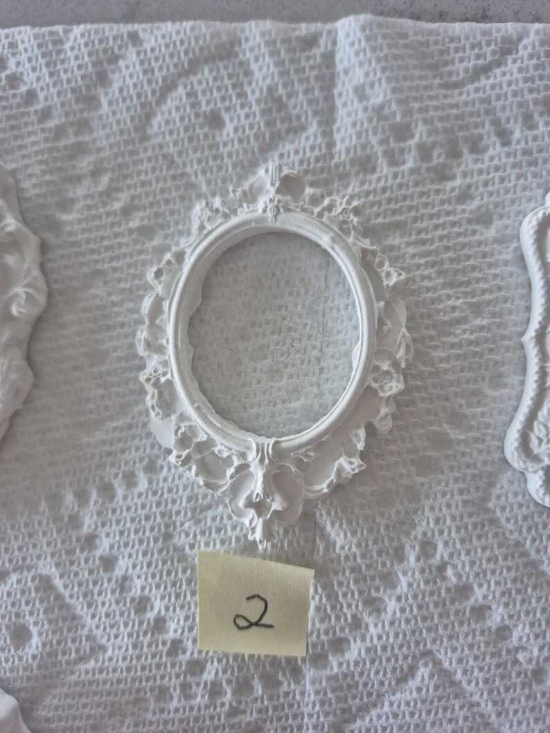 May include: White oval-shaped picture frame with ornate detailing. The frame has a decorative floral design around the edges. A small yellow sticky note with the number "2" is visible below the frame. The frame is on a white textured surface.