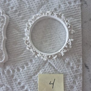 May include: A white, circular, decorative frame with intricate floral detailing. The frame is placed on a white, textured surface. A small, yellow sticky note with the number "4" written in black ink is also visible.