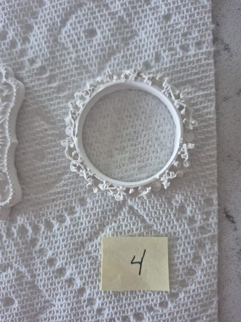 May include: A white, circular, decorative frame with intricate floral detailing. The frame is placed on a white, textured surface. A small, yellow sticky note with the number "4" written in black ink is also visible.