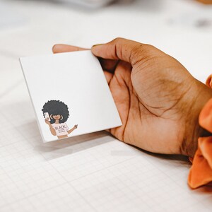 Może przedstawiać: Biały notes z kresk&oacute;wkową ilustracją czarnej kobiety z afrykańskim warkoczem, kt&oacute;ra nosi r&oacute;żową koszulkę z napisem "Black Girl Magic" i trzyma smartfona.