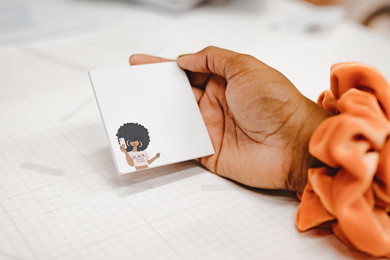 Może przedstawiać: Biały notes z kresk&oacute;wkową ilustracją czarnej kobiety z afrykańskim warkoczem, kt&oacute;ra nosi r&oacute;żową koszulkę z napisem "Black Girl Magic" i trzyma smartfona.