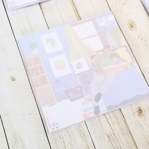 May include: Square notepad with an illustration of a parent and child reading in bed. The image has a soft, pastel color palette with a bookshelf, dresser, and framed artwork in the background. A soccer ball is in the corner.