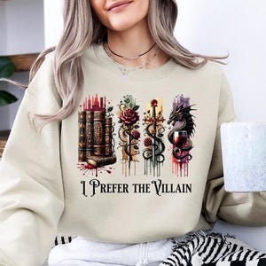 May include: Beige sweatshirt with a watercolor design featuring books, roses, swords, a dragon, and a wine glass. The text "I Prefer the Villain" is printed below the design. The person is holding a white and blue mug.