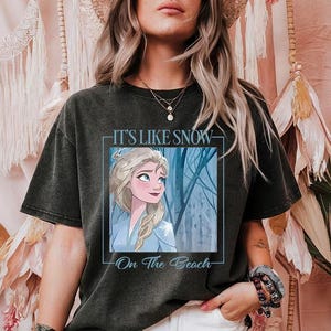 May include: A black t-shirt with a graphic of a cartoon character with blonde hair and blue eyes. The text on the shirt reads "It's Like Snow On The Beach".