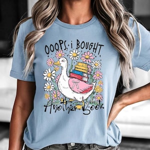 May include: Pale blue t-shirt with a graphic of a goose laden with books, encircled by flowers. The text reads "OOOPS, I BOUGHT Another Book". A lighthearted design for book lovers.