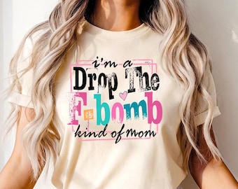 May include: Cream-colored t-shirt with the phrase "I'm a Drop The F-bomb kind of mom" in a colorful, playful font. The text is framed in a pink rectangle, with a small heart graphic. The shirt has short sleeves.