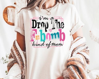 May include: Cream-colored t-shirt with the text "I'm a Drop The F-bomb kind of mom" in black, pink, teal, and orange. The graphic is inside a pink and black rectangular border. The shirt has short sleeves.