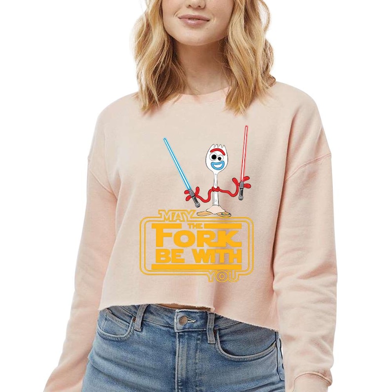 May include: A light peach-colored cropped sweatshirt featuring a graphic of a white plastic spork character with red pipe cleaner arms holding a blue and a red lightsaber. Below the character, the yellow text reads MAY THE FORK BE WITH YOU.