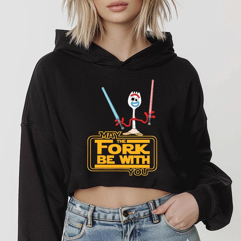 Puede incluir: Una sudadera negra corta con capucha que muestra un gr&aacute;fico de un tenedor de pl&aacute;stico blanco con brazos de limpiapipas rojos sosteniendo un sable de luz azul y otro rojo. El texto amarillo dice: MAY THE FORK BE WITH YOU.