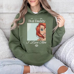 May include: Olive green hooded sweatshirt with the text "But Daddy I Love Him" and a cartoon image of a red-haired character. The sweatshirt has a front pocket and a drawstring hood.