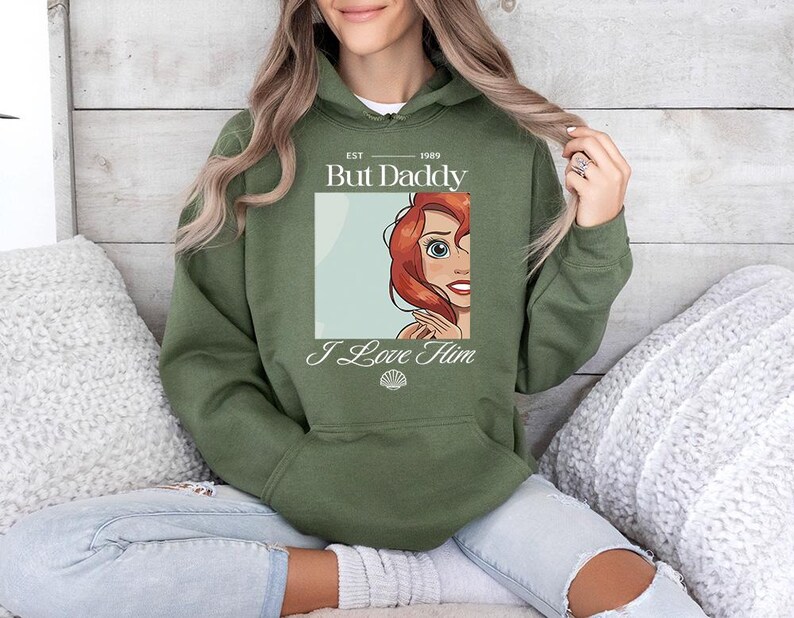May include: Olive green hooded sweatshirt with the text "But Daddy I Love Him" and a cartoon image of a red-haired character. The sweatshirt has a front pocket and a drawstring hood.