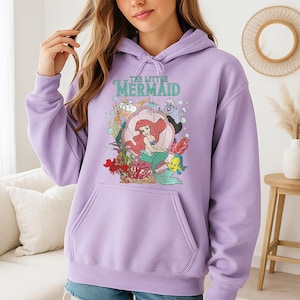 May include: A light purple hooded sweatshirt featuring a graphic print of The Little Mermaid characters including Ariel, King Triton, Ursula, Sebastian, and Flounder. The design includes a large pink seashell, coral, and bubbles on the front.