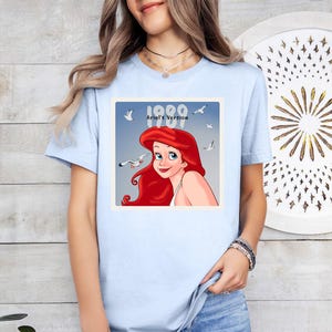 May include: Pale blue t-shirt with a graphic of Ariel from The Little Mermaid, with the text "1989 Ariel's Version." The image shows Ariel with red hair, blue eyes, and seagulls. A casual, everyday wear tee.
