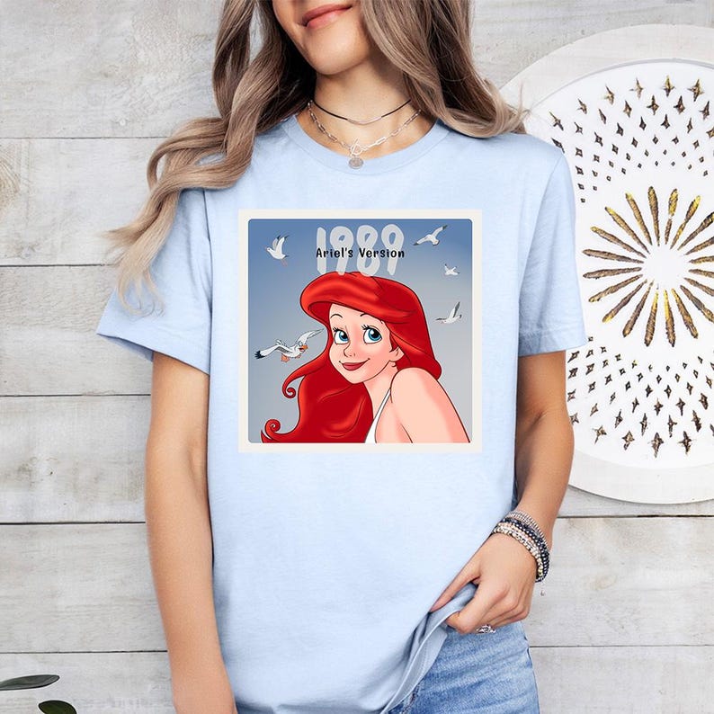 May include: Pale blue t-shirt with a graphic of Ariel from The Little Mermaid, with the text "1989 Ariel's Version." The image shows Ariel with red hair, blue eyes, and seagulls. A casual, everyday wear tee.