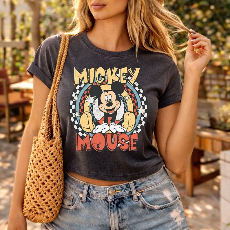 Pu&ograve; includere: Una t-shirt grigio antracite a maniche corte con una grafica di Mickey Mouse centrata all'interno di un design circolare. La stampa include il testo MICKEY MOUSE in giallo e rosso, circondato da un bordo a scacchi, stelle e accenti blu.