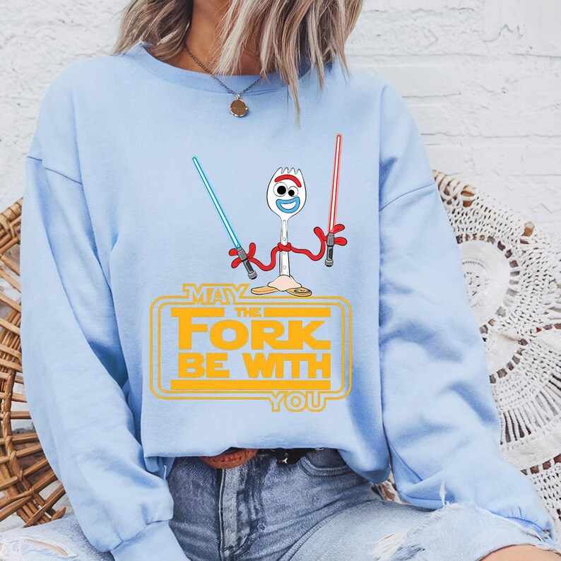 May include: A light blue crewneck sweatshirt featuring a graphic of a white plastic spork character with googly eyes holding a blue and a red lightsaber. Below the character, the yellow text reads: MAY THE FORK BE WITH YOU.