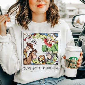 Puede incluir: Sudadera blanca con un gr&aacute;fico colorido de personajes de Toy Story dentro de un borde negro. El texto "YOU'VE GOT A FRIEND IN ME" est&aacute; impreso debajo. La sudadera la lleva una persona.