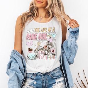 Può includere: Canotta bianca con una grafica colorata che recita "THE LIFE OF A PARK GIRL". La grafica include immagini di cartoni animati di un castello, popcorn, una tazza da tè e un gelato di Topolino. Indossata con una giacca di jeans.