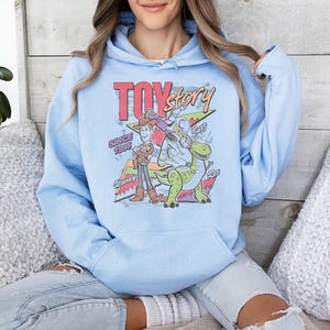 May include: A light blue hooded sweatshirt featuring a graphic print of Toy Story characters Woody, Buzz Lightyear, and Rex. The design includes the text Toy Story, Since 1995, Woody, Buzz, and Rex in a colorful, retro-inspired font and layout.
