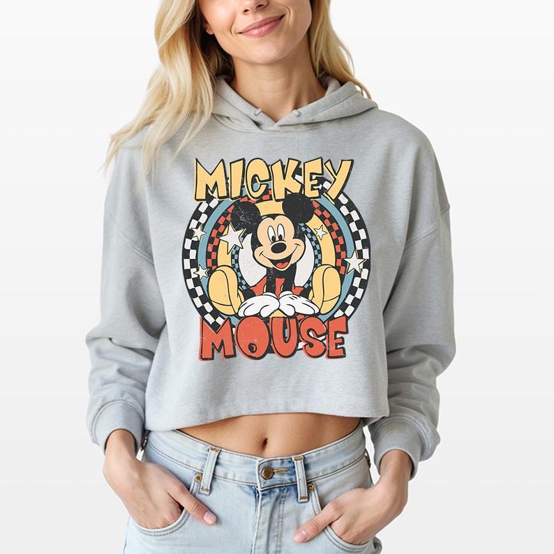 May include: A light gray cropped hoodie featuring a graphic print of Mickey Mouse. The design includes the text "MICKEY MOUSE" in yellow and red, surrounded by a black and white checkered circle, colorful concentric rings, and white stars.