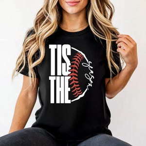 May include: Black t-shirt with the words "TIS THE SEASON" in white block letters. A baseball graphic with red stitching and the word "season" in script is on the right side. The shirt is a casual style.