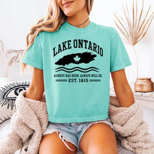 May include: A light turquoise short-sleeve t-shirt featuring black text that reads LAKE ONTARIO, a black silhouette of the lake with a white maple leaf, wavy lines, and the text ALWAYS HAS BEEN. ALWAYS WILL BE. EST. 1615.