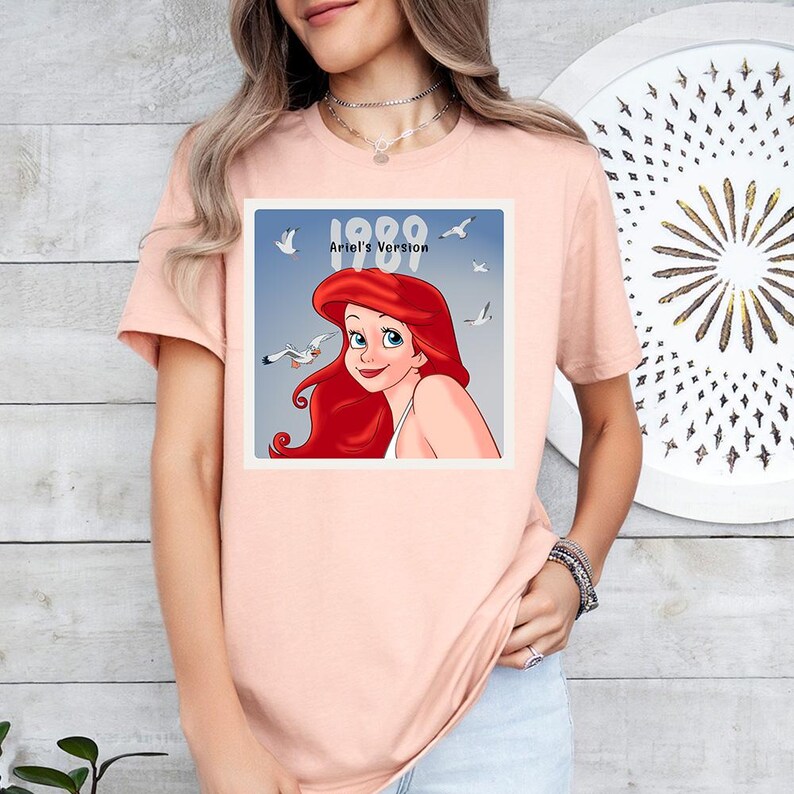 May include: A pale pink t-shirt with a square graphic of Ariel from The Little Mermaid, with the text "1989 Ariel's Version". The graphic shows Ariel, seagulls, and a blue sky background.