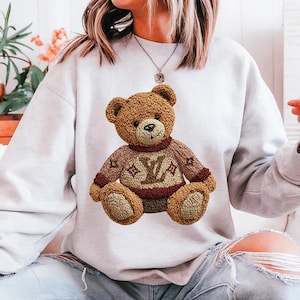 May include: A light gray sweatshirt featuring a brown teddy bear with a sweater. The teddy bear's sweater is brown with a burgundy collar and the letter "V" and a star design. The teddy bear is sitting on the sweatshirt.