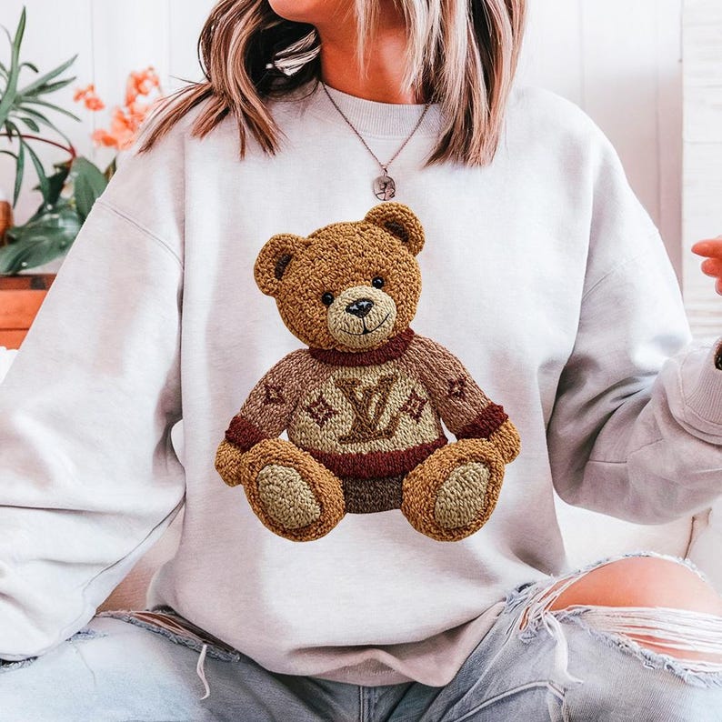 May include: A light gray sweatshirt featuring a brown teddy bear with a sweater. The teddy bear's sweater is brown with a burgundy collar and the letter "V" and a star design. The teddy bear is sitting on the sweatshirt.