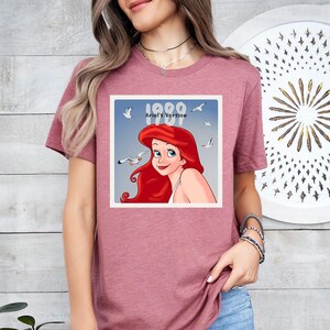 May include: A mauve t-shirt with a graphic of Ariel from The Little Mermaid. The graphic is in a square frame with the text "1989 Ariel's Version" and seagulls. Ariel has red hair and blue eyes.