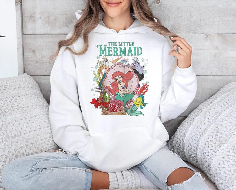 May include: A white hooded sweatshirt featuring a graphic print of The Little Mermaid characters including Ariel, King Triton, Ursula, Sebastian, and Flounder. The design includes the text The Little Mermaid in teal lettering above the characters.