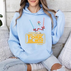 Op de afbeelding: Een lichtblauwe hoodie met een afbeelding van een witte plastic spork met rode pijpenrager-armen die een blauw en een rood lichtzwaard vasthoudt. Onder het figuur staat de gele tekst: MAY THE FORK BE WITH YOU.