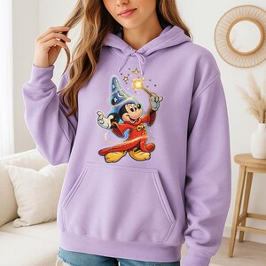 May include: A light purple hoodie featuring a large graphic of Mickey Mouse dressed as a wizard. Mickey is wearing a blue wizard hat with stars and a red robe, holding a wand with a glowing star. The hoodie has a front pocket and a drawstring hood.