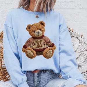 May include: A light blue sweatshirt featuring a detailed embroidered teddy bear wearing a brown sweater with a logo design. The bear has a brown body and is sitting upright. The sweatshirt has long sleeves.