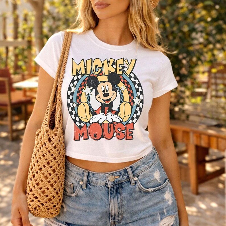 Pu&ograve; includere: Una t-shirt bianca a maniche corte con una grafica di Mickey Mouse centrata all'interno di un design circolare. La grafica include un bordo a scacchi, stelle e il testo MICKEY MOUSE in lettere grassetto e usurate nei colori giallo e rosso.