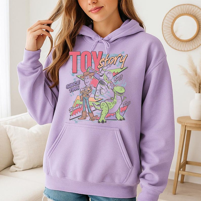 May include: A light purple hooded sweatshirt featuring a graphic print of Toy Story characters Woody, Buzz Lightyear, and Rex. The design includes the text Toy Story, Since 1995, Woody, Buzz, and Rex, set against colorful geometric shapes and stars.
