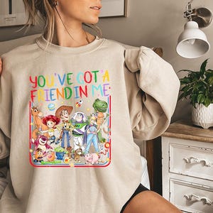 以下が含まれることがあります： 様々なアニメーションのおもちゃのキャラクターが描かれたカラフルなグラフィックプリントが特徴のベージュのクルーネックトレーナー。キャラクターの上には「YOU'VE GOT A FRIEND IN ME」というテキストが多色の文字でプリントされ、星や惑星のアイコンが添えられています。