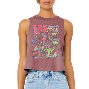 Puede incluir: Un top corto sin mangas de color malva con un estampado gr&aacute;fico de los personajes de Toy Story: Woody, Buzz Lightyear y Rex. El dise&ntilde;o incluye el texto "TOY story" en rosa y amarillo, "SINCE 1995" en azul y formas geom&eacute;tricas coloridas de fondo.