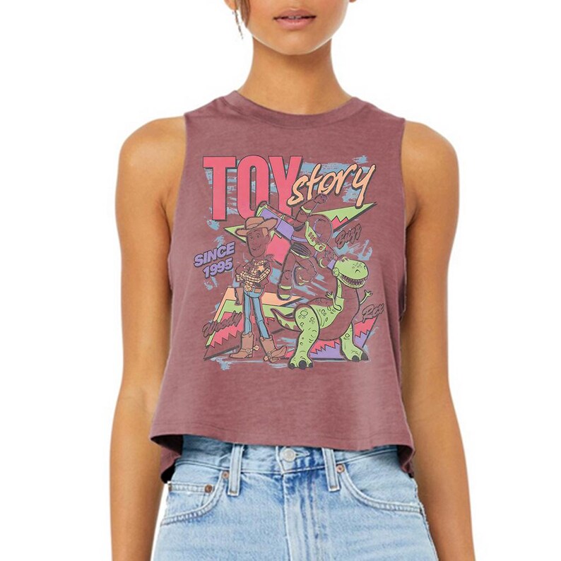 Puede incluir: Un top corto sin mangas de color malva con un estampado gr&aacute;fico de los personajes de Toy Story: Woody, Buzz Lightyear y Rex. El dise&ntilde;o incluye el texto "TOY story" en rosa y amarillo, "SINCE 1995" en azul y formas geom&eacute;tricas coloridas de fondo.