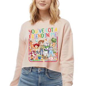 Peut inclure: Un sweat-shirt court de couleur p&ecirc;che clair avec une impression graphique color&eacute;e repr&eacute;sentant divers personnages de jouets anim&eacute;s. Le texte "YOU'VE GOT A FRIEND IN ME" est imprim&eacute; en lettres multicolores au-dessus des personnages, incluant un cow-boy, un ranger de l'espace et une cow-girl.