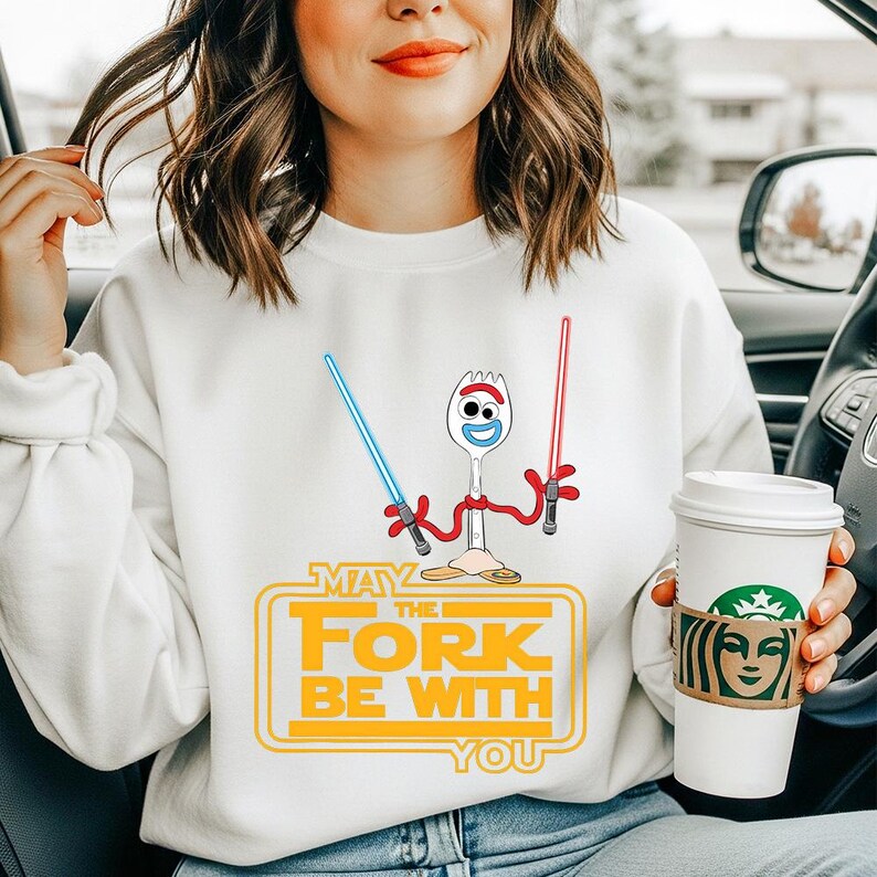 May include: A white crewneck sweatshirt featuring a graphic of a spork character holding a blue lightsaber in one hand and a red lightsaber in the other. Below the character, the text reads MAY THE FORK BE WITH YOU in yellow block letters.