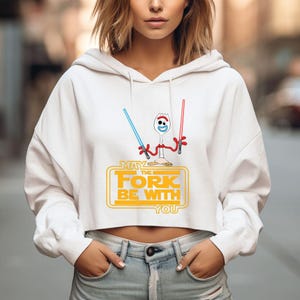 Puede incluir: Una sudadera corta blanca con capucha que presenta un gr&aacute;fico de un personaje tipo tenedor sosteniendo un sable de luz azul y otro rojo. Debajo, el texto MAY THE FORK BE WITH YOU aparece en letras amarillas. Tiene mangas largas y capucha.