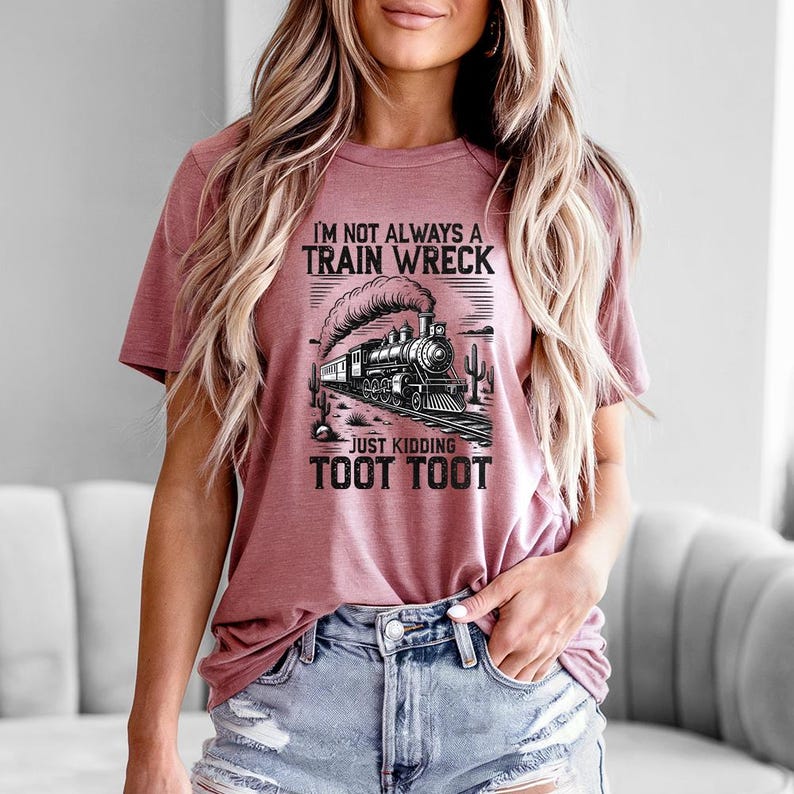 May include: Dusty rose t-shirt with a black and white graphic of a train and the text "I'M NOT ALWAYS A TRAIN WRECK JUST KIDDING TOOT TOOT". The shirt has short sleeves and a crew neck.