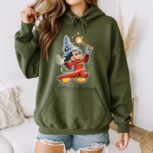 May include: Olive green hooded sweatshirt featuring a graphic of Mickey Mouse dressed as a wizard. Mickey is wearing a blue wizard hat with stars and a red robe, holding a wand with a glowing star. The sweatshirt has a front pocket and a drawstring hood.