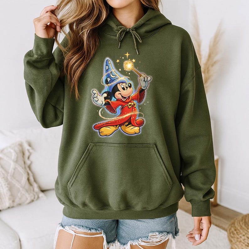 May include: Olive green hooded sweatshirt featuring a graphic of Mickey Mouse dressed as a wizard. Mickey is wearing a blue wizard hat with stars and a red robe, holding a wand with a glowing star. The sweatshirt has a front pocket and a drawstring hood.