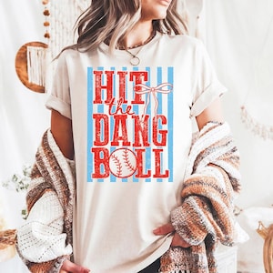 May include: A cream-coloured t-shirt featuring the text "HIT the DANG BALL" in red, with a baseball graphic. The text is set against a light blue striped background. A red bow is above the text. The shirt is short-sleeved.