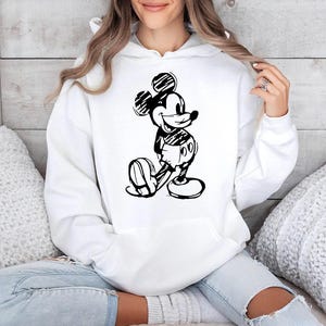 May include: White hooded sweatshirt featuring a black and white sketch of Mickey Mouse. The cartoon character is depicted in a classic pose. The sweatshirt has a front pocket and a drawstring hood. The design is a simple, yet iconic, graphic.