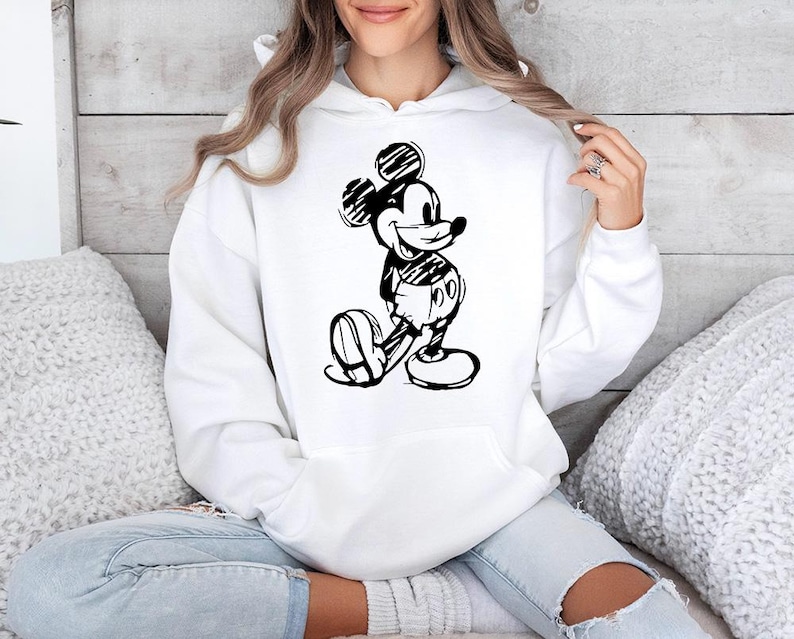 May include: White hooded sweatshirt featuring a black and white sketch of Mickey Mouse. The cartoon character is depicted in a classic pose. The sweatshirt has a front pocket and a drawstring hood. The design is a simple, yet iconic, graphic.